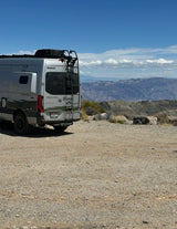 Winnebago Revel RV rental parked on gravel with ladder & roof rack, overlooking a vast mountain landscape under a blue sky.