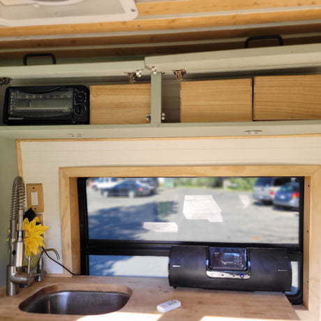 Cozy RV rental interior with kitchen, overhead storage, and parking lot view. Features toaster oven, sink, sound system.