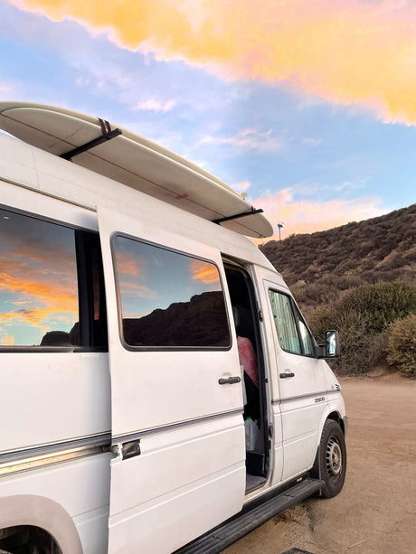 White RV rental, surfboards ready, enjoys a stunning sunset by a hillside.