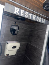 Compact RV rental restroom: dark tiled walls, round mirror, and a white, motion-sensor trash can.