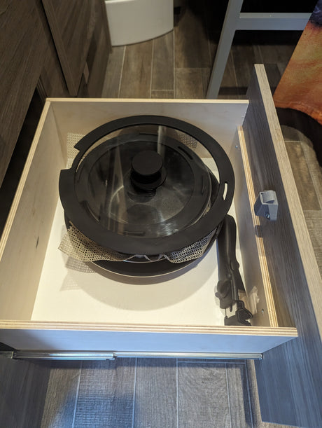 An open light-wood drawer in an RV rental reveals nesting cookware (pot, pan, lid) and a detachable handle, secured for travel.