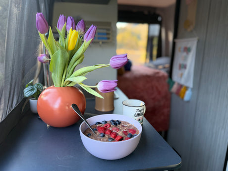 Cozy morning in an RV rental: Enjoy oatmeal with berries, fresh tulips, and a "Happy Camper" mug amidst nature.