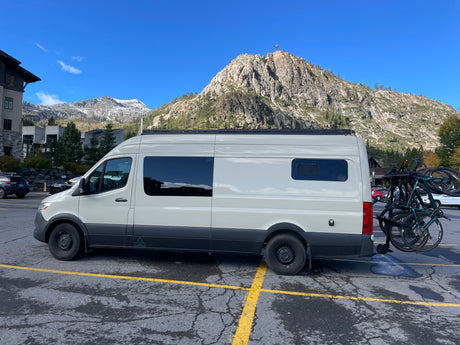 White **rv rental** van with bicycles on the back, parked in a mountain resort lot, ready for outdoor adventures.