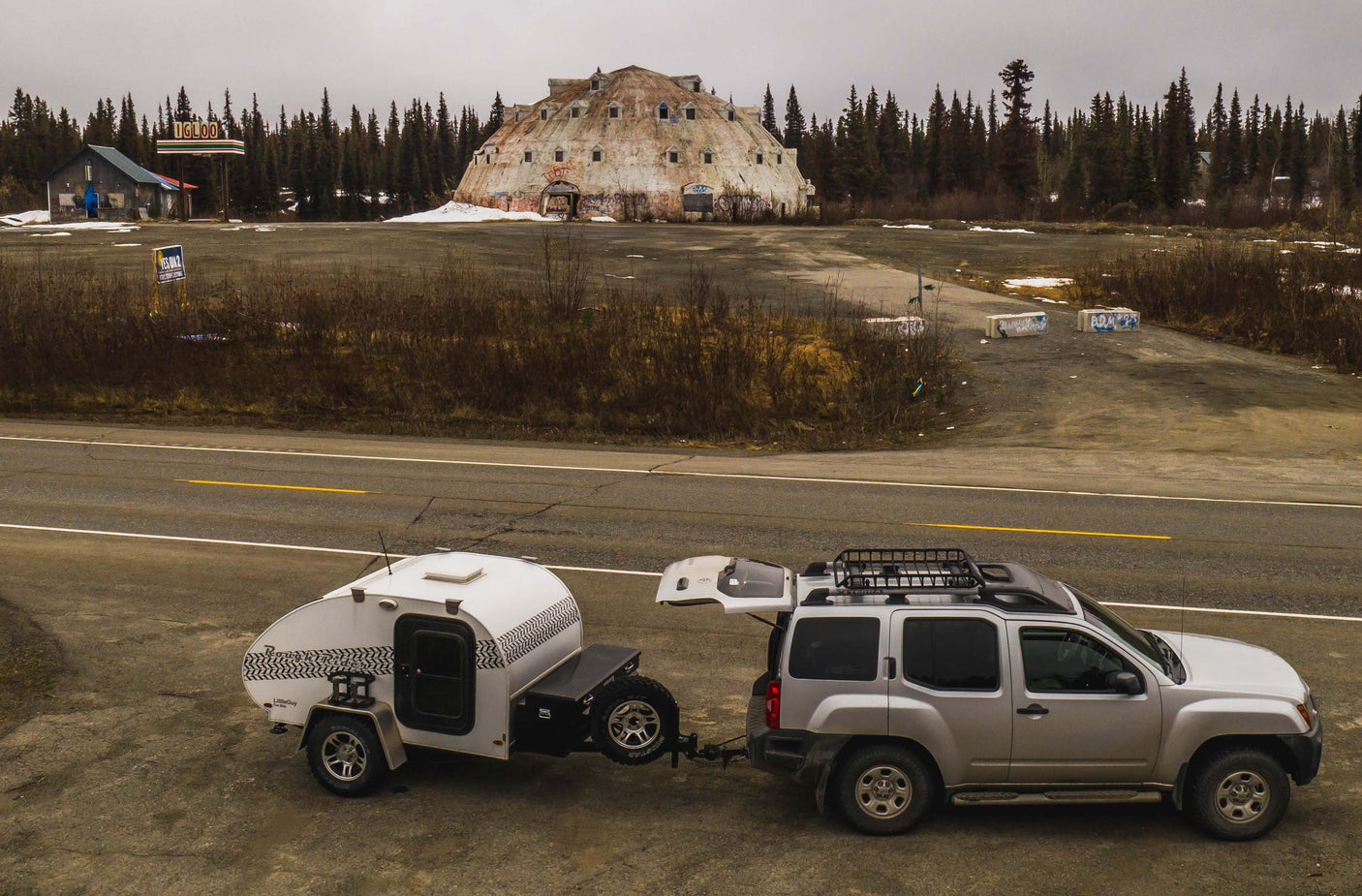An RV rental setup: a silver SUV tows a teardrop camper. Parked by the road, with the abandoned Igloo Hotel in the background.
