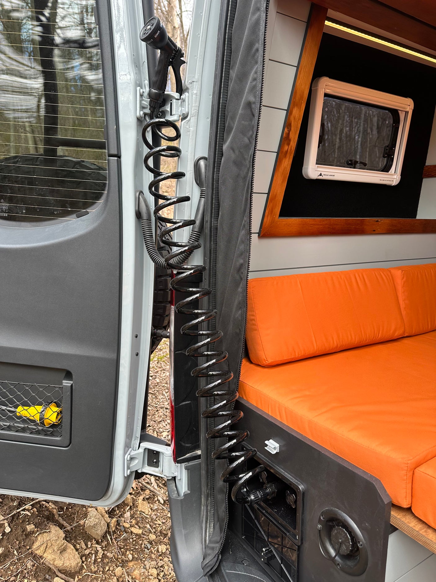 Back view of an RV rental: open door, a black coiled outdoor shower hose, and a cozy interior with orange cushions.
