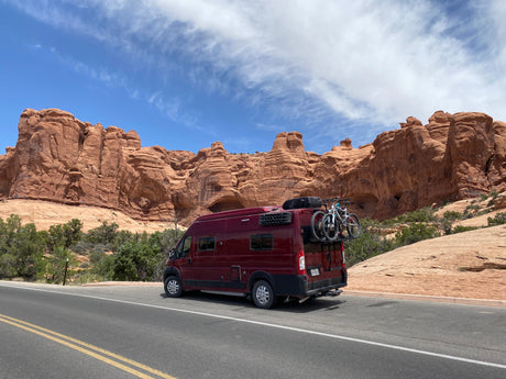 Maroon RV rental with bikes parked on a road, flanked by towering red rock formations under a blue sky with wispy clouds.