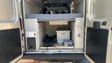 View into an **rv rental** van's organized rear. Features a raised bed, pull-out storage, and essential utilities like outlets/water spigot.