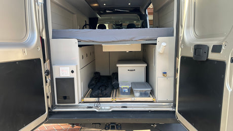 View into an **rv rental** van's organized rear. Features a raised bed, pull-out storage, and essential utilities like outlets/water spigot.