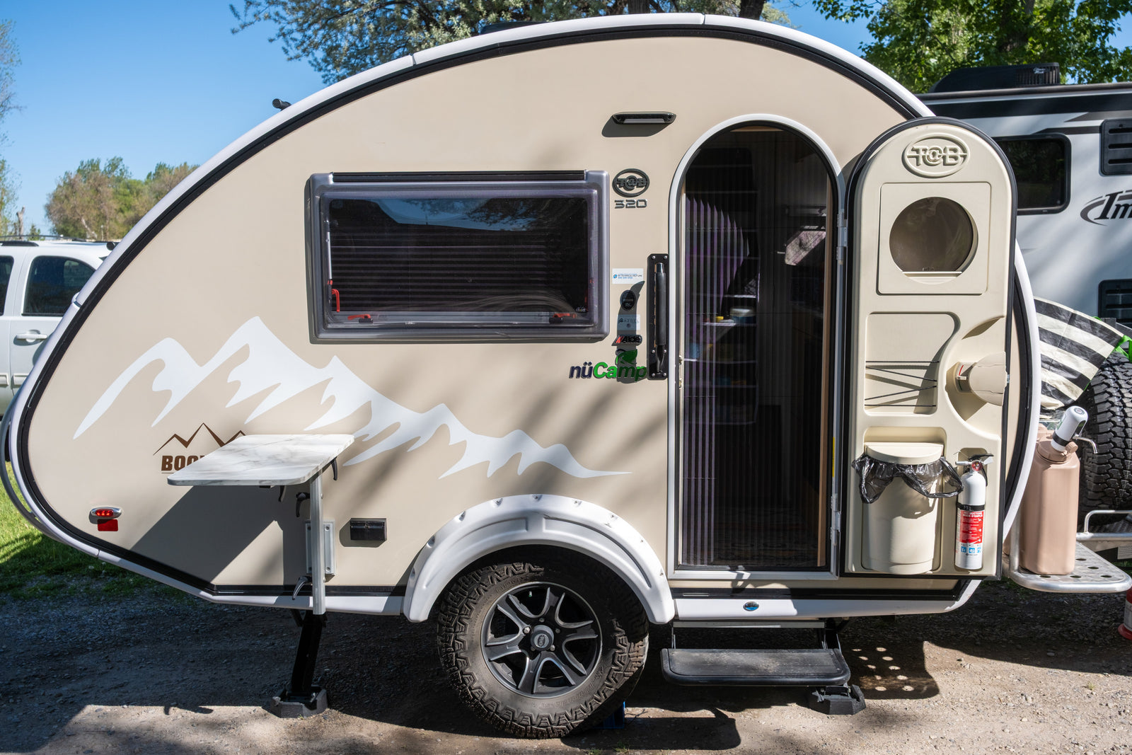 This beige nuCamp T@B 320 teardrop camper is ideal for an RV rental. Features a side table, open door, & mountain graphics.