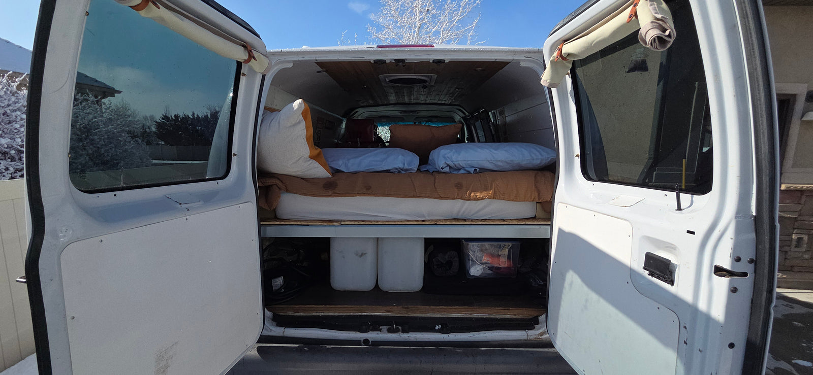RV rental van interior with a bed, under-bed storage, and open rear doors revealing a snowy landscape outside.
