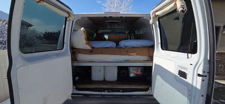 RV rental van interior with a bed, under-bed storage, and open rear doors revealing a snowy landscape outside.