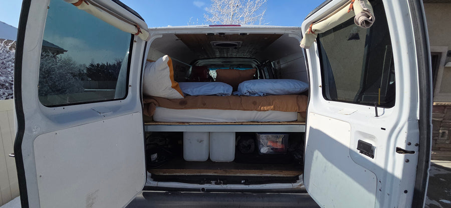RV rental van interior with a bed, under-bed storage, and open rear doors revealing a snowy landscape outside.
