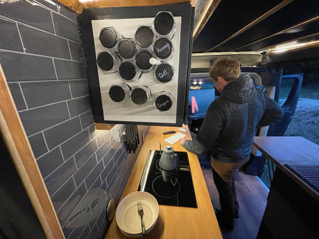Modern rv rental interior: kitchen with magnetic spice rack, induction cooktop, and a person utilizing a computer workstation.