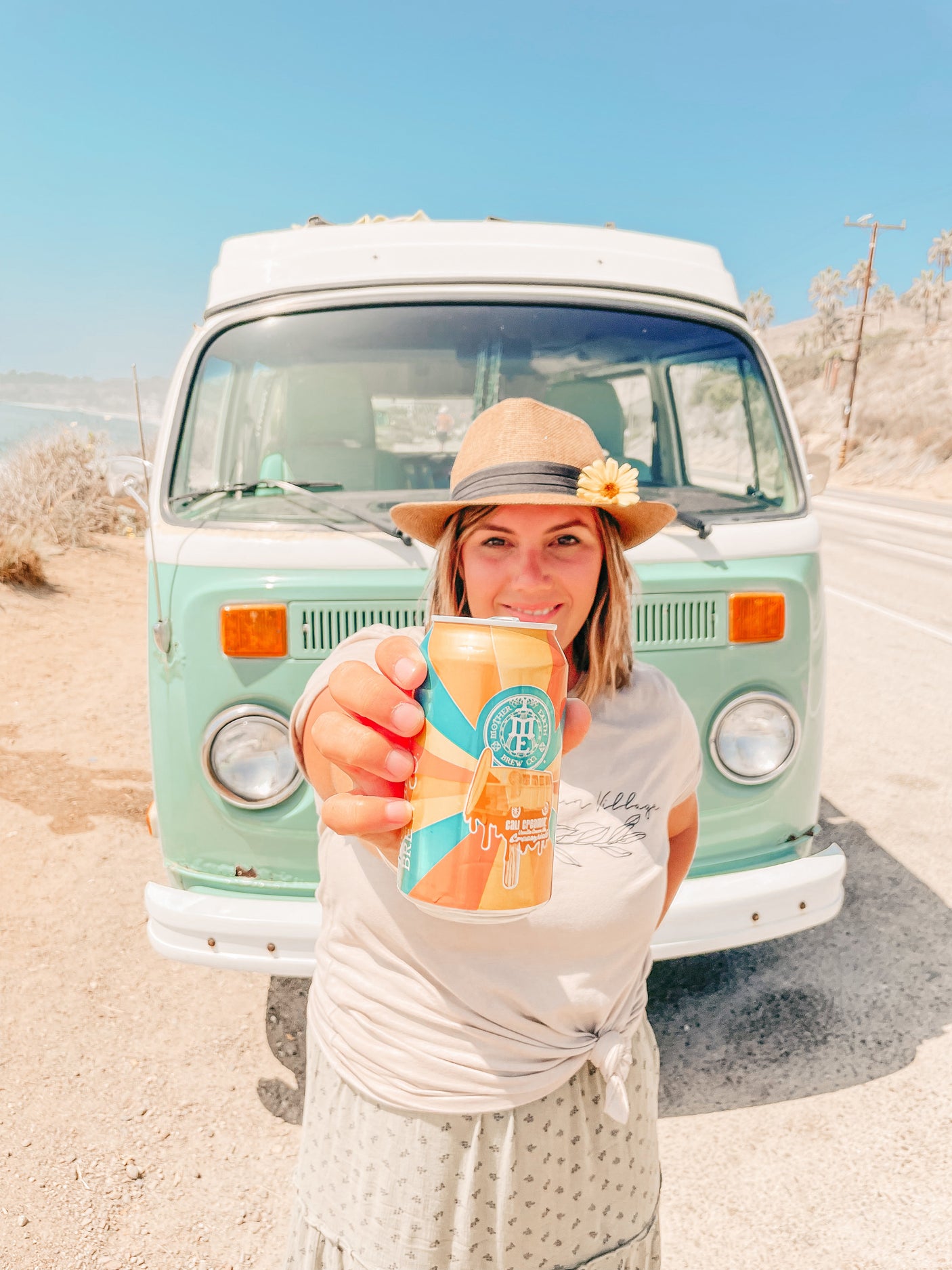 Smiling woman holding a can, in front of a vintage VW camper van on a sunny coastal road. Dream RV rental adventure!