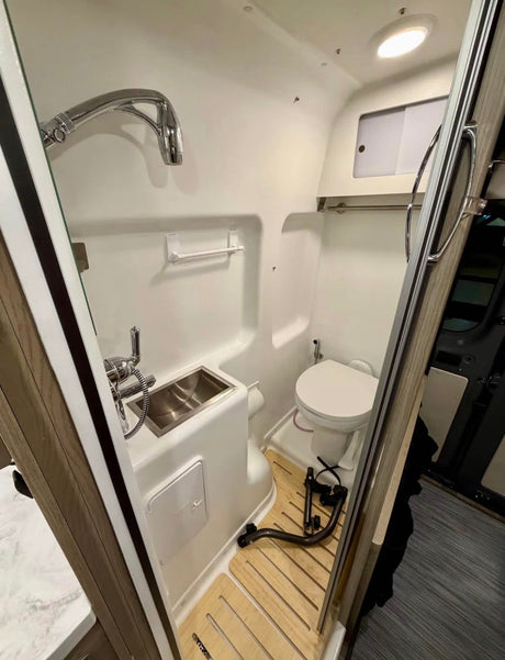 Inside an RV rental: a compact wet bath with shower, small sink, and toilet. White walls, wooden floor.