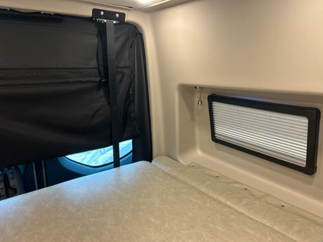 Inside an rv rental: a light-colored bed, a black privacy curtain over a window, and a small window with blinds.