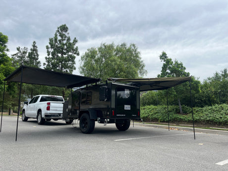RV rental: White pickup truck and dark green camper trailer with extended awnings set up in a parking lot under a cloudy sky.