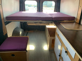 Inside a compact RV rental: a comfortable purple bed, kitchen counter, and storage unit for travel.
