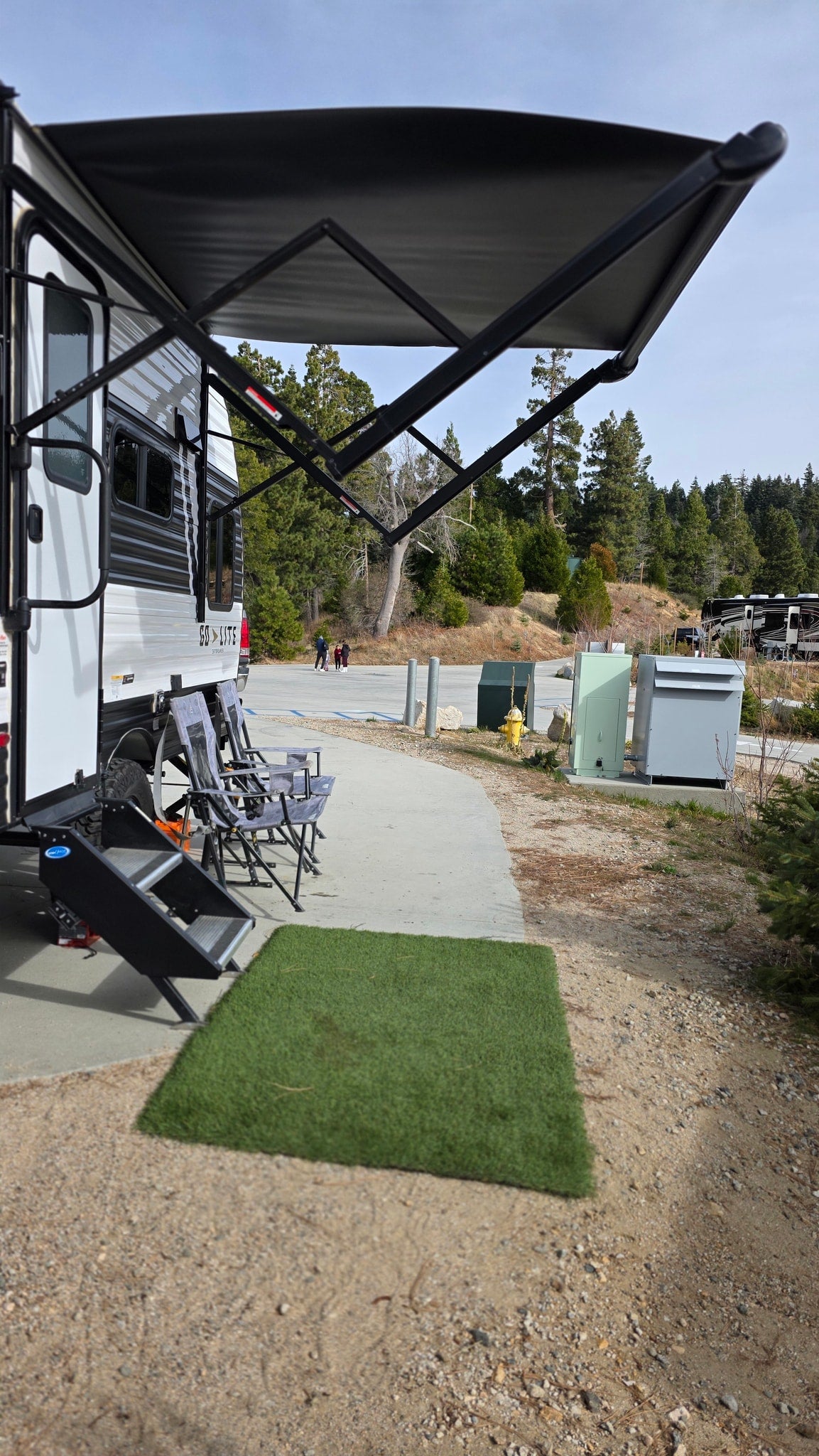 RV rental at a scenic campsite with an extended awning, chairs, and steps ready for relaxation amidst pine trees. — photo 9