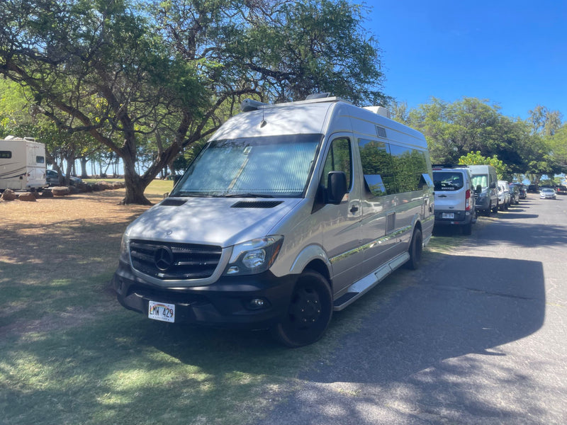 Hawaii RV rental: Silver Mercedes Sprinter van parked in a shaded campground with other vans under a clear sky.|Dusky sky over a gray camper van RV rental with bright fireworks in the distance.|RV rental sleeping area: a neatly made bed with white sheets & pillows — photo 1