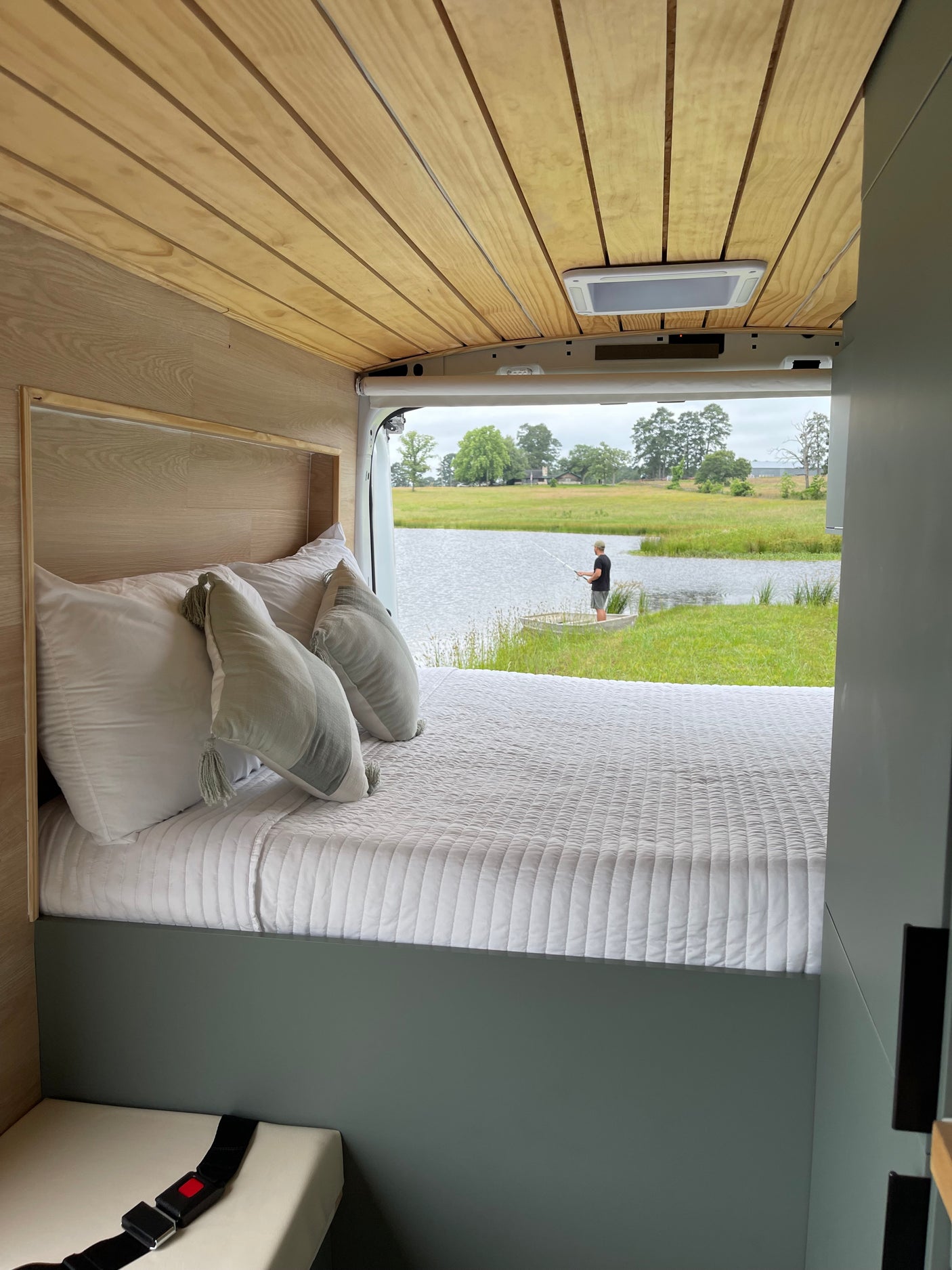 Cozy RV rental interior with a comfortable bed. View through open back: a person fishing in a lake surrounded by green landscape.