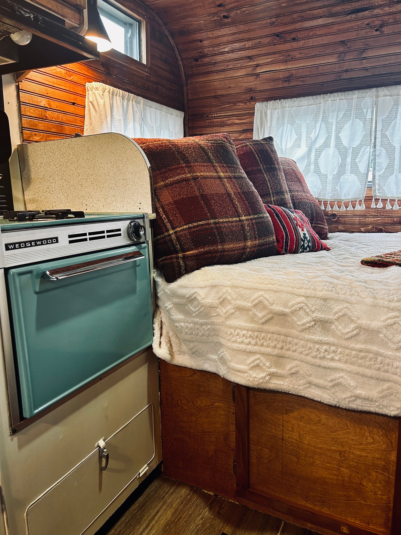 Cozy RV rental interior featuring a vintage Wedgewood stove, plush bed with plaid pillows, and warm wood paneling.