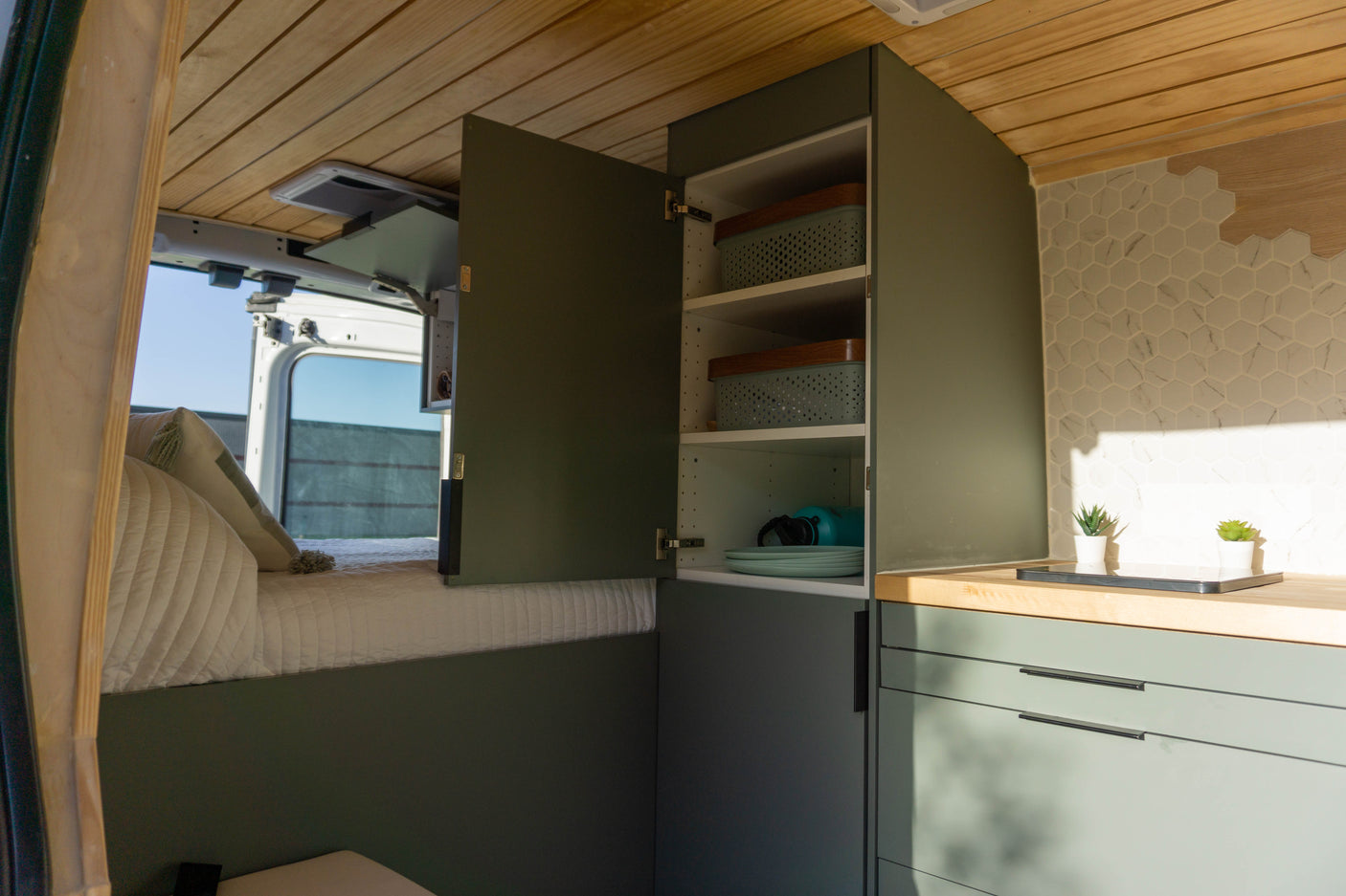 Inside an RV rental: a modern, custom-built camper van featuring a cozy bed, olive-green cabinetry, a wood counter, and tile backsplash.