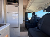Compact RV rental interior with kitchen, desk, laptop, and driver's area, offering a scenic beach view.
