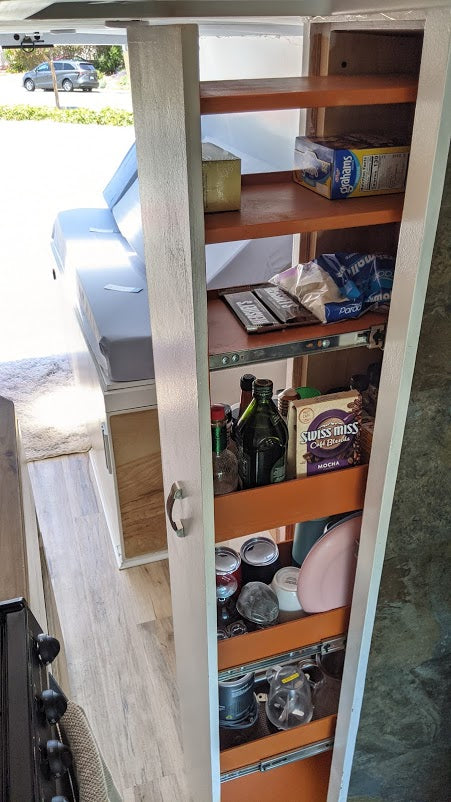 RV rental interior: A compact, organized pull-out pantry stocked with snacks and kitchen essentials next to a cozy bed.