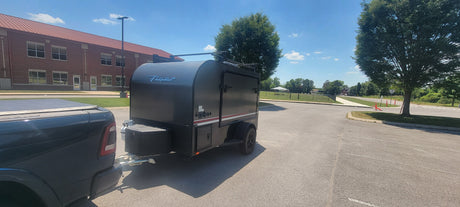 A black Flyer camping trailer, ideal for rv rental adventures, hitched to a truck in a parking lot. Ready for outdoor travel.