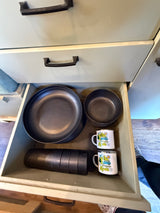 RV rental kitchen drawer, neatly stocked with black plates, cups, and enamel "Adventure Awaits" mugs for travels.