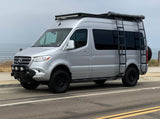 This silver Mercedes Sprinter RV rental boasts off-road tires, a rugged bumper, and a roof rack with ladder.