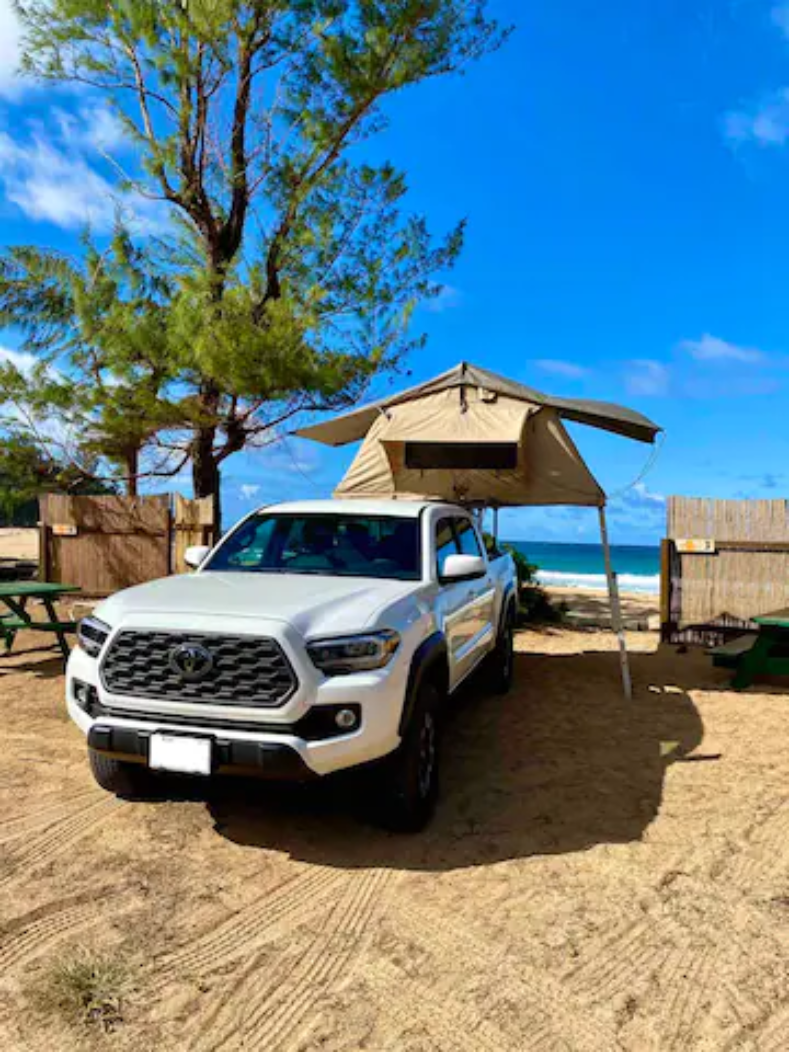 White Toyota Tacoma RV rental, with a rooftop tent and awning, parked on a sandy beach. Trees & ocean in view for camping.