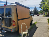 Tan camper van, ready for RV rental adventures, with its large side awning deployed, parked on a sunny street.