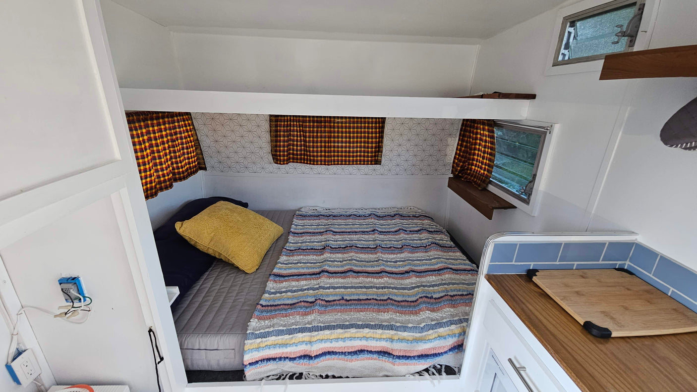 Inside a compact RV rental: a bed with striped blanket, plaid curtains, and a small kitchenette area.