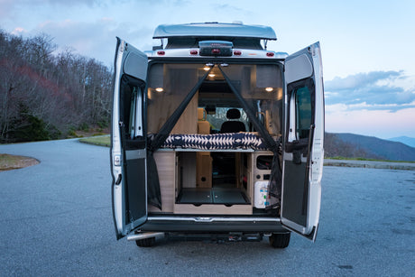 Camper van RV rental with open rear doors showing bed and interior setup, parked on a road with mountains.