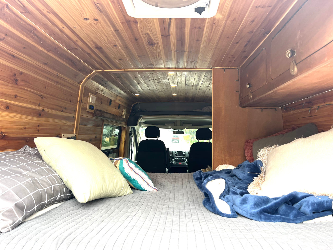Warm wood-paneled RV rental interior with a comfy bed in the foreground, looking towards the vehicle's driver cabin.