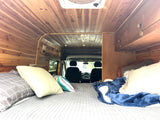 Warm wood-paneled RV rental interior with a comfy bed in the foreground, looking towards the vehicle's driver cabin.