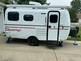 White Scamp travel trailer RV rental, parked on concrete with awning, AC, and leveling jacks.