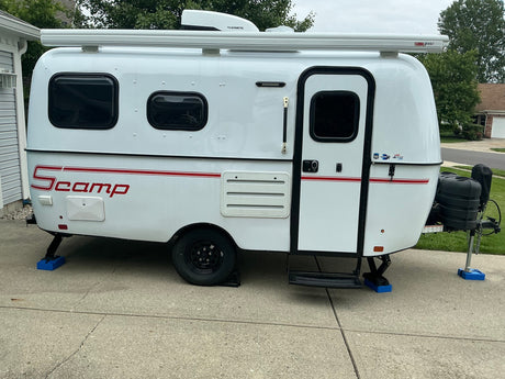 White Scamp travel trailer RV rental, parked on concrete with awning, AC, and leveling jacks.