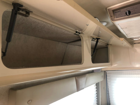 Overhead cabinets with soft-lined interiors and gas struts in an RV rental. An AC unit and window with pleated blinds are also seen.