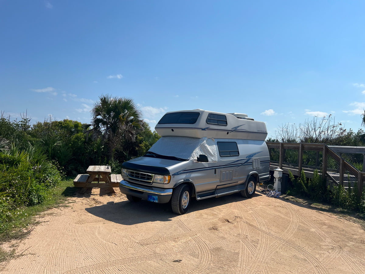Silver RV rental parked on a sandy campsite with a picnic table, surrounded by lush greenery and palm trees under a clear blue sky. — photo 9