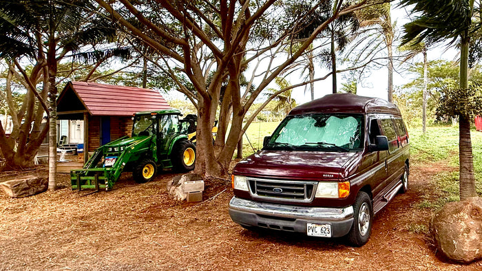 Maroon van, likely an RV rental, with a green tractor, shed, and trees. Dirt ground. #rvrental