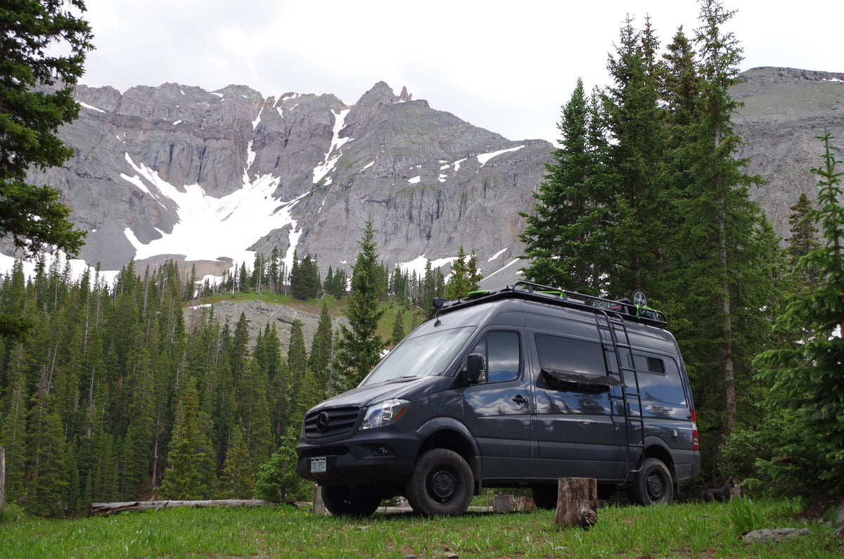 Dark gray RV rental van parked in a scenic mountain meadow with snow-capped peaks and evergreen trees in the background.