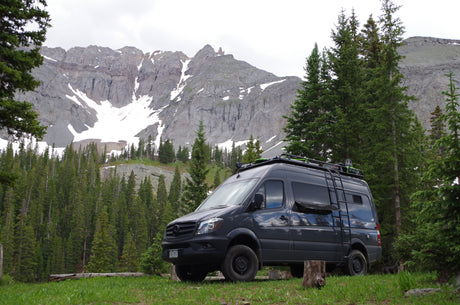 Dark gray RV rental van parked in a scenic mountain meadow with snow-capped peaks and evergreen trees in the background.