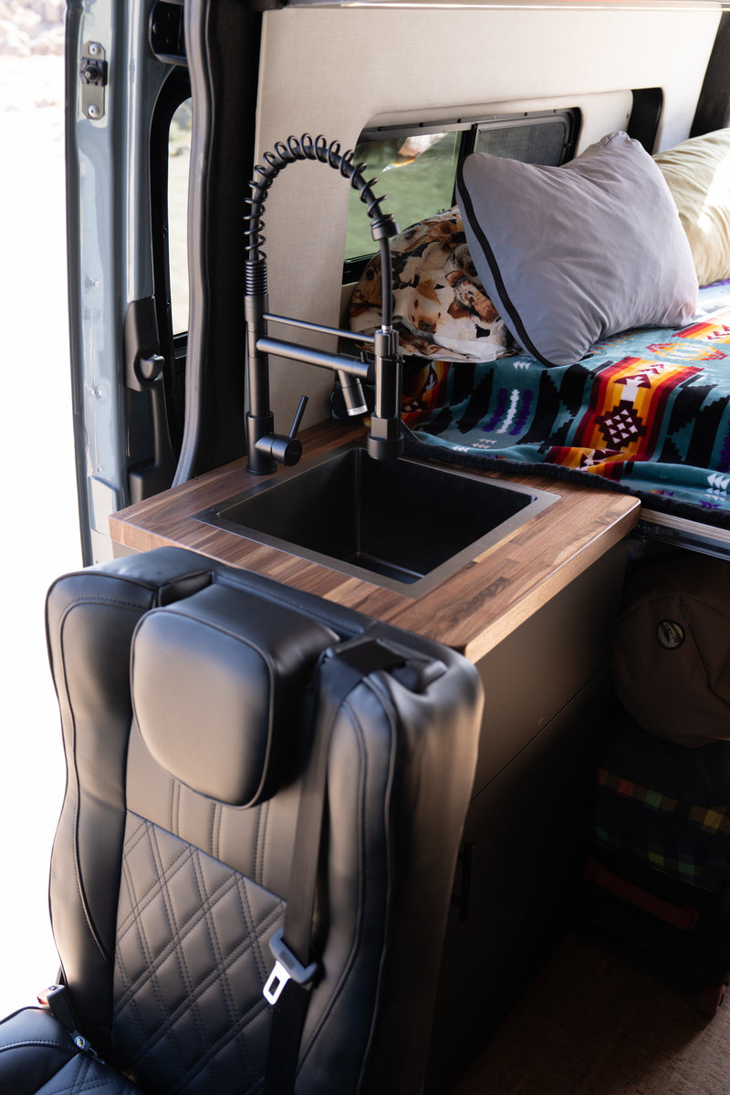 Experience an RV rental! Modern van interior featuring a sleek black sink, wooden counter, and quilted leather seat near the cozy bed. — photo 8