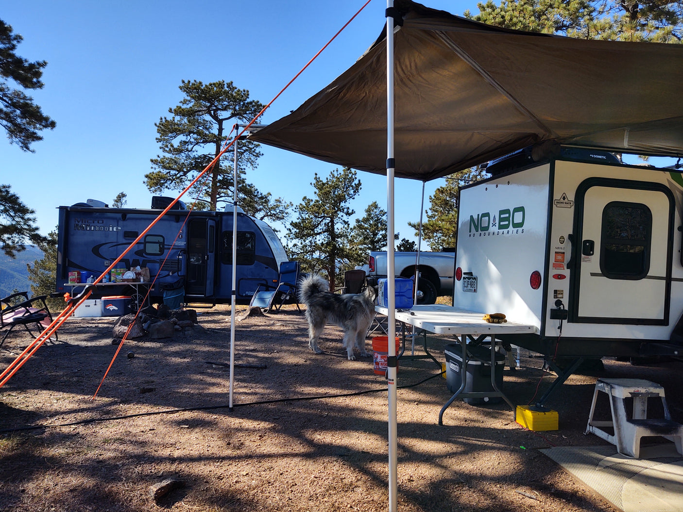 Sunny rv rental campsite: A white No Boundaries camper and a blue Micro Minnie are set up with an awning, tables, and a dog.
