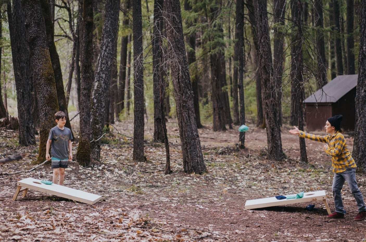 Two people enjoy an outdoor cornhole game in a forest, a perfect scene for your RV rental adventure. A beanbag is mid-air.