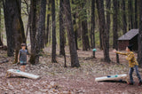 Two people enjoy an outdoor cornhole game in a forest, a perfect scene for your RV rental adventure. A beanbag is mid-air.