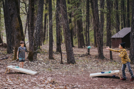 Two people enjoy an outdoor cornhole game in a forest, a perfect scene for your RV rental adventure. A beanbag is mid-air.